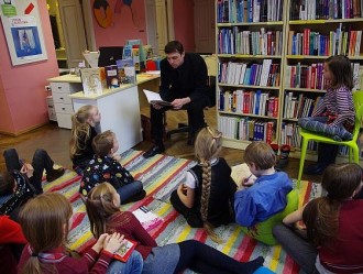 lapsed istuvad värvilisel triibulisel vaibal ja kuulvad kuidas neile üks mees raamatut ette loeb