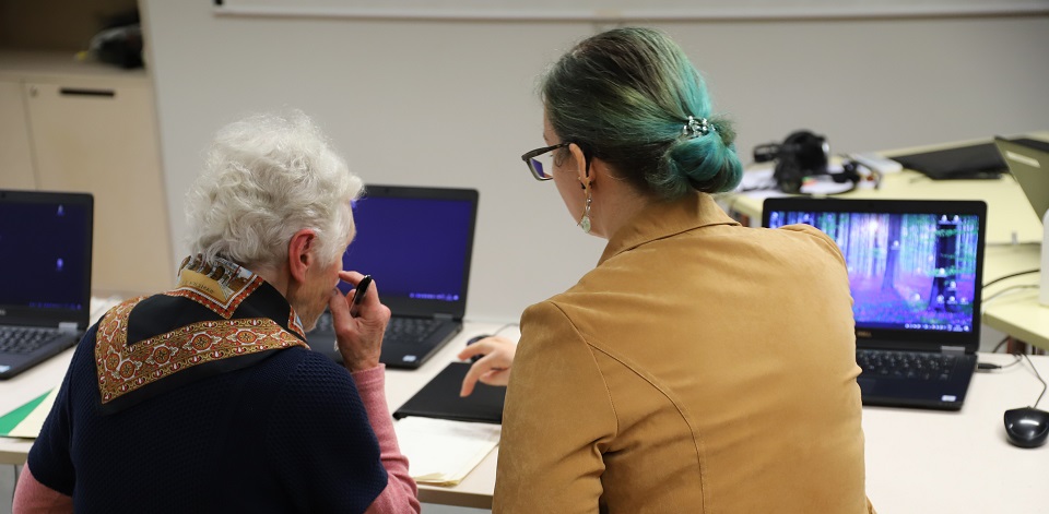 Raamatukogu töötaja juhendab vanemat daami sülearvuti kasutamises. Mõlemad on tagant vaates.
